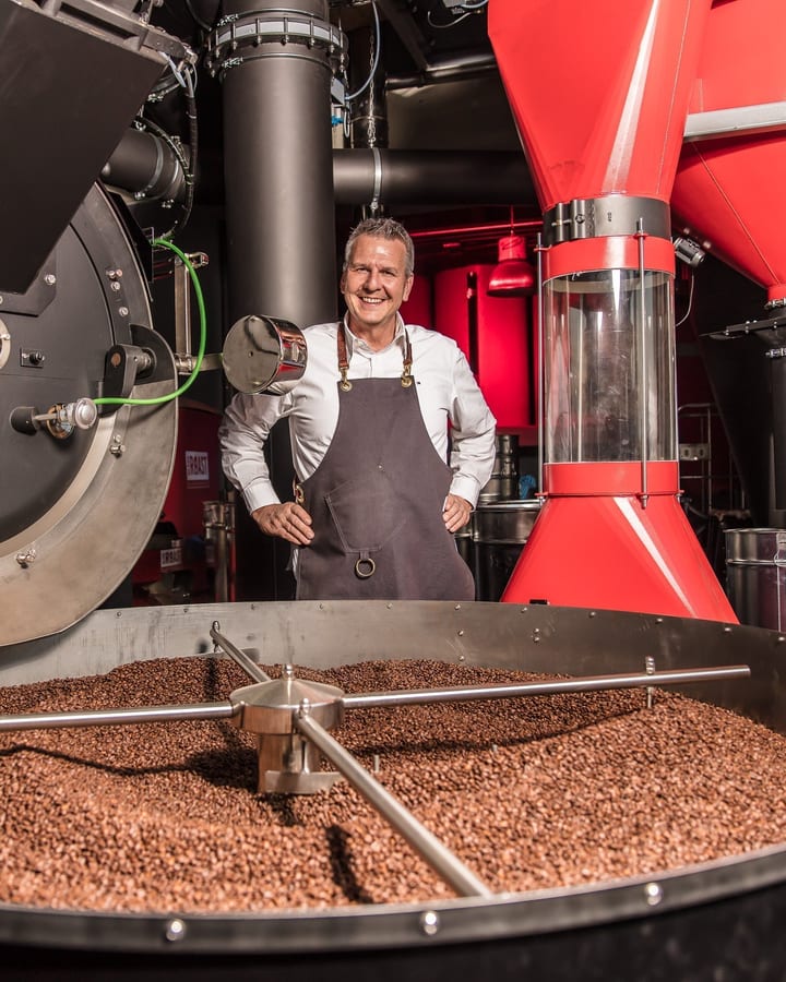 Roaster in an apron smiling between the drum roaster and a red silo, with the cooling tray full of coffee beans in front