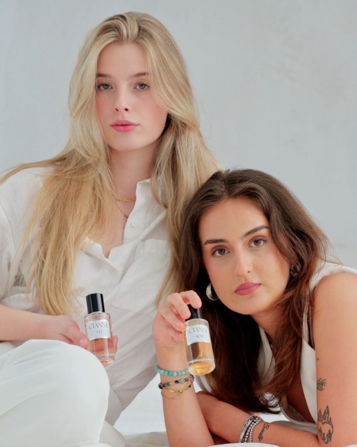 Two young women in white lying on a bed holding perfume bottles.