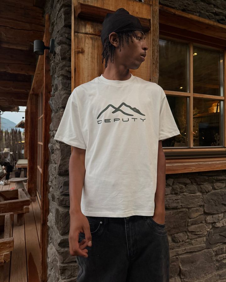 Young man in a white Deputy T-shirt with mountain print in front of a wooden chalet