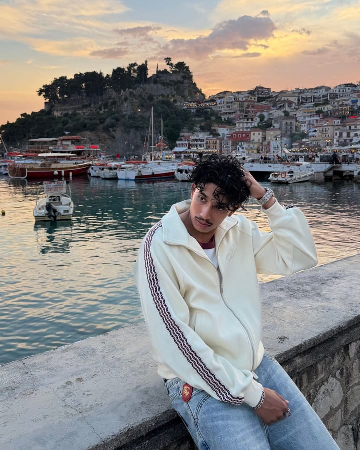 Man in a cream track jacket leaning on a harbour wall with boats and the old town in evening light behind him