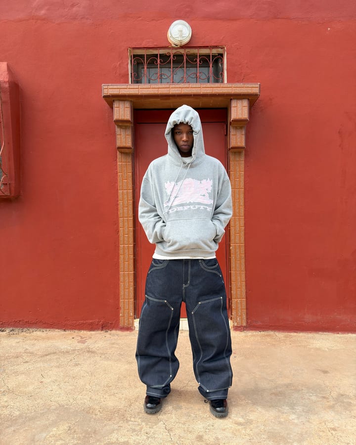 Mann in grauem Hoodie und weiter dunkler Jeans steht vor einer roten Hauswand mit Holztür
