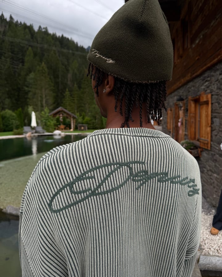 Rückenansicht: grün gestreifter Strickpullover mit Deputy-Schriftzug und dunkle Mütze vor Bergsee und Chalet