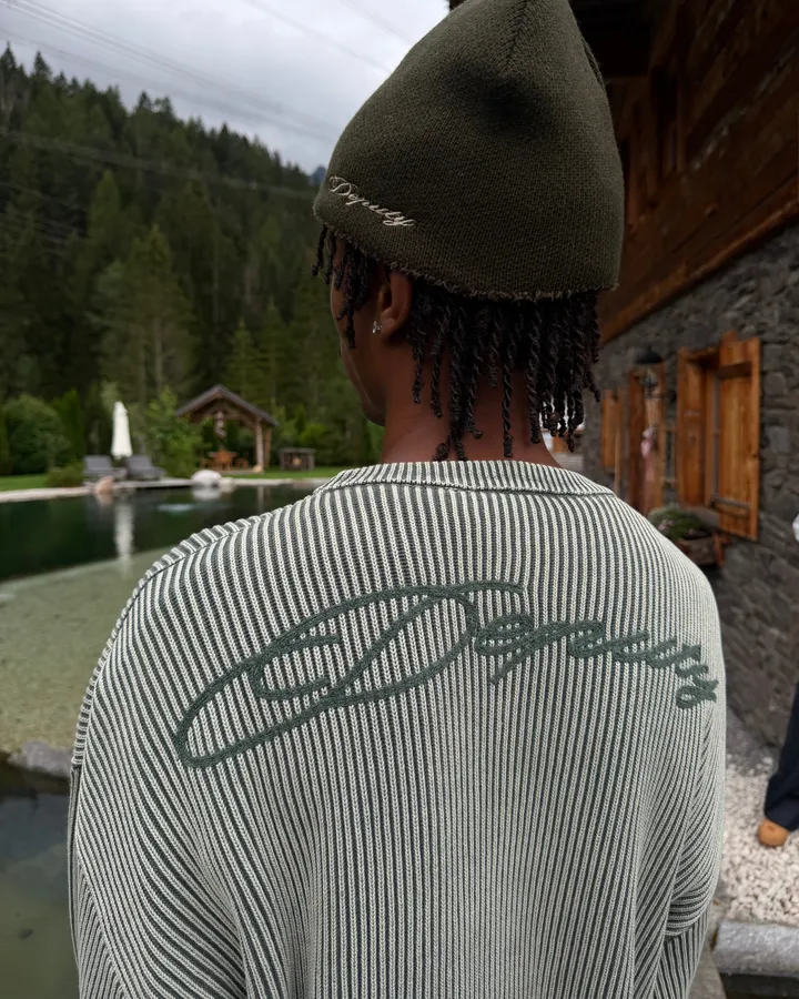 Back view of a green striped knit jumper with Deputy lettering and a dark beanie in front of a mountain lake and chalet