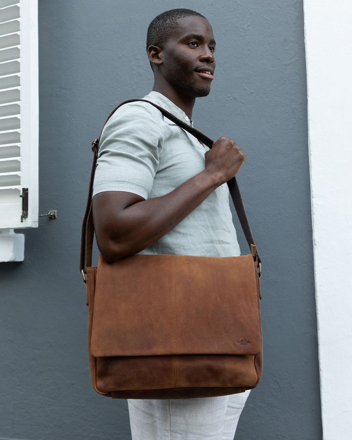 Man carrying a brown leather messenger bag in front of a grey wall
