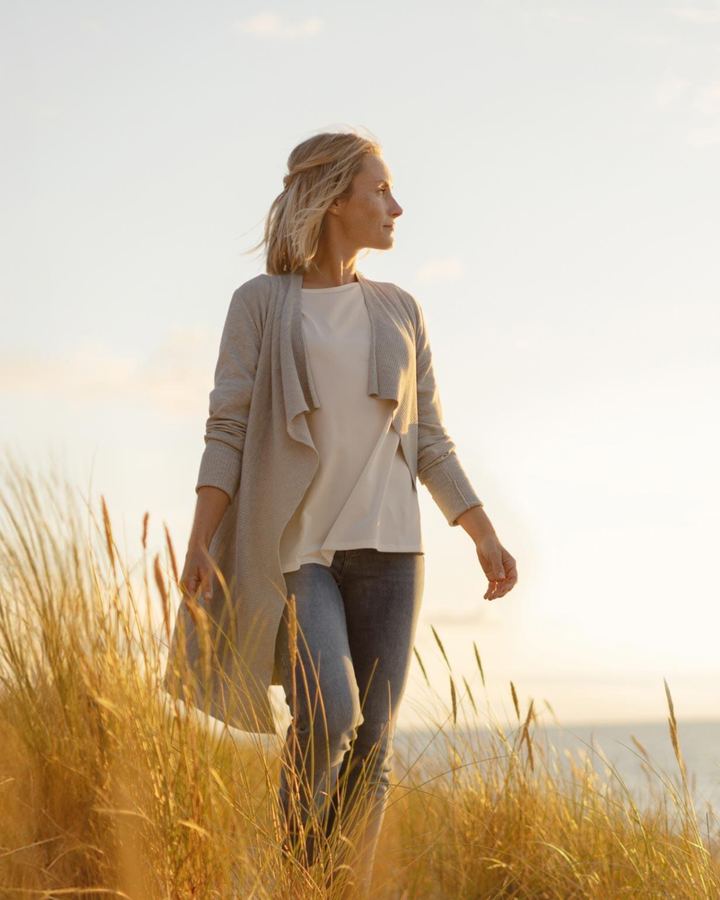 Woman in a cardigan walking through dune grass by the sea in evening light.