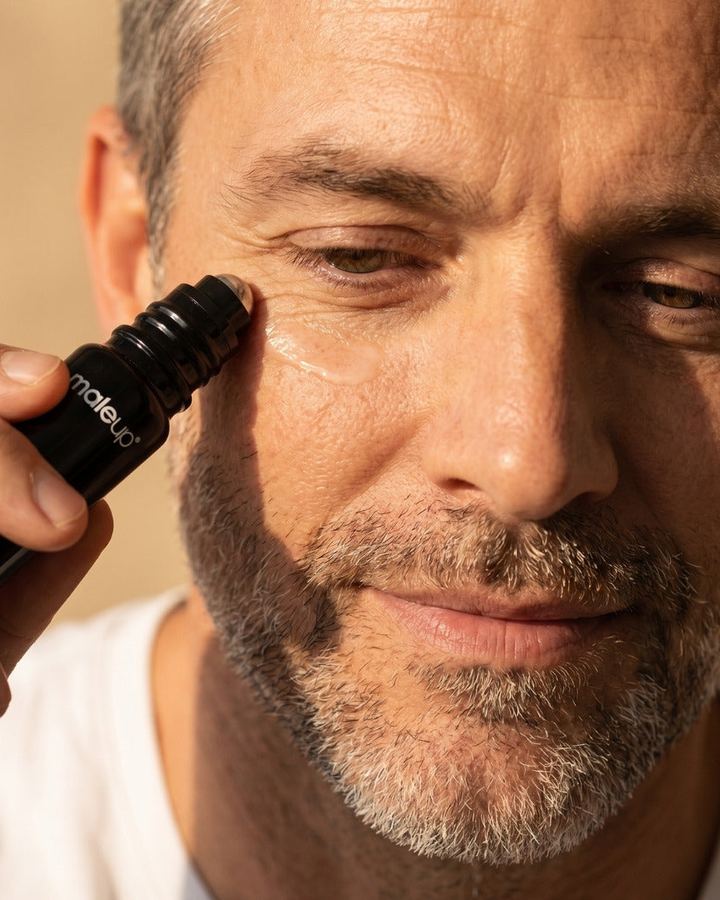 Mann mit Bart trägt mit einem Roll-on-Serum Pflege unter dem Auge auf, warmes Seitenlicht.