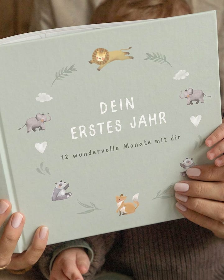 Hände halten ein grünes Babytagebuch mit Tierillustrationen und dem Titel Dein erstes Jahr