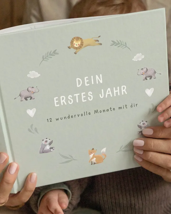 Hände halten ein grünes Babytagebuch mit Tierillustrationen und dem Titel Dein erstes Jahr