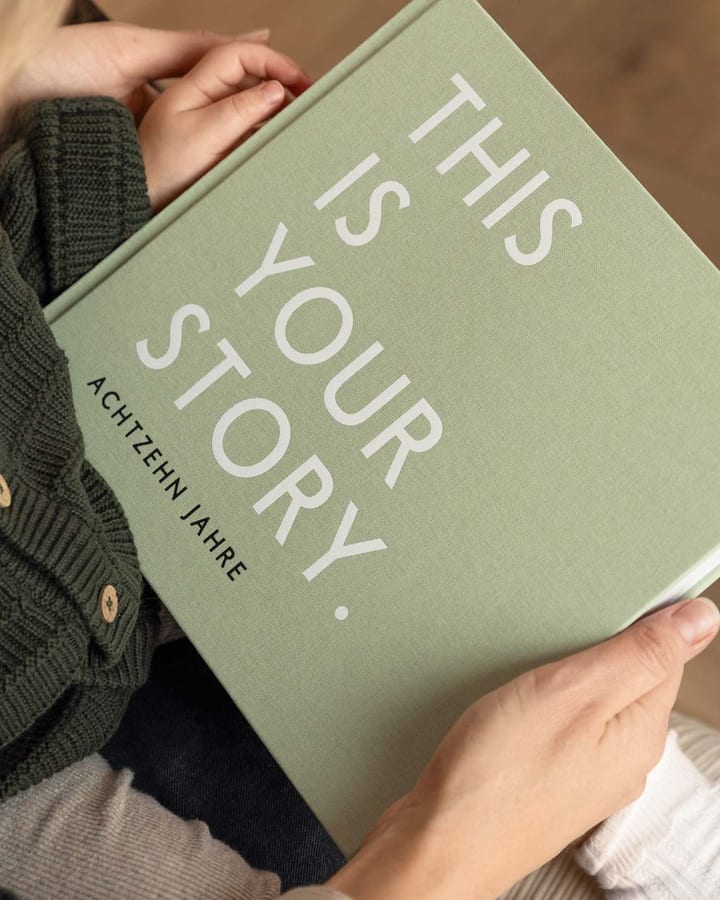 Hände halten ein salbeigrünes Erinnerungsbuch mit der Aufschrift This is your story