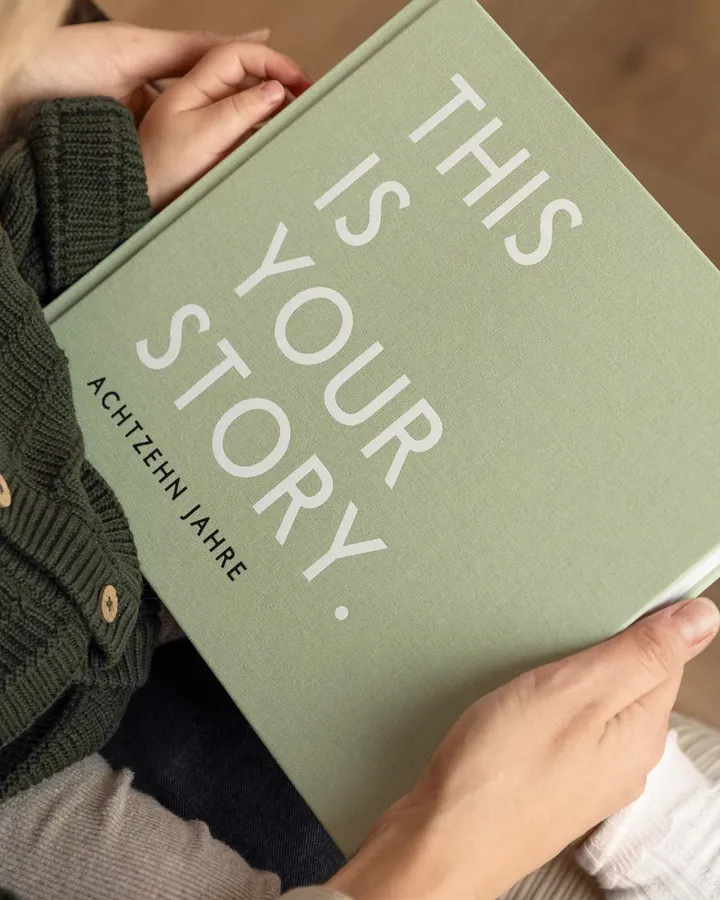 Hände halten ein salbeigrünes Erinnerungsbuch mit der Aufschrift This is your story