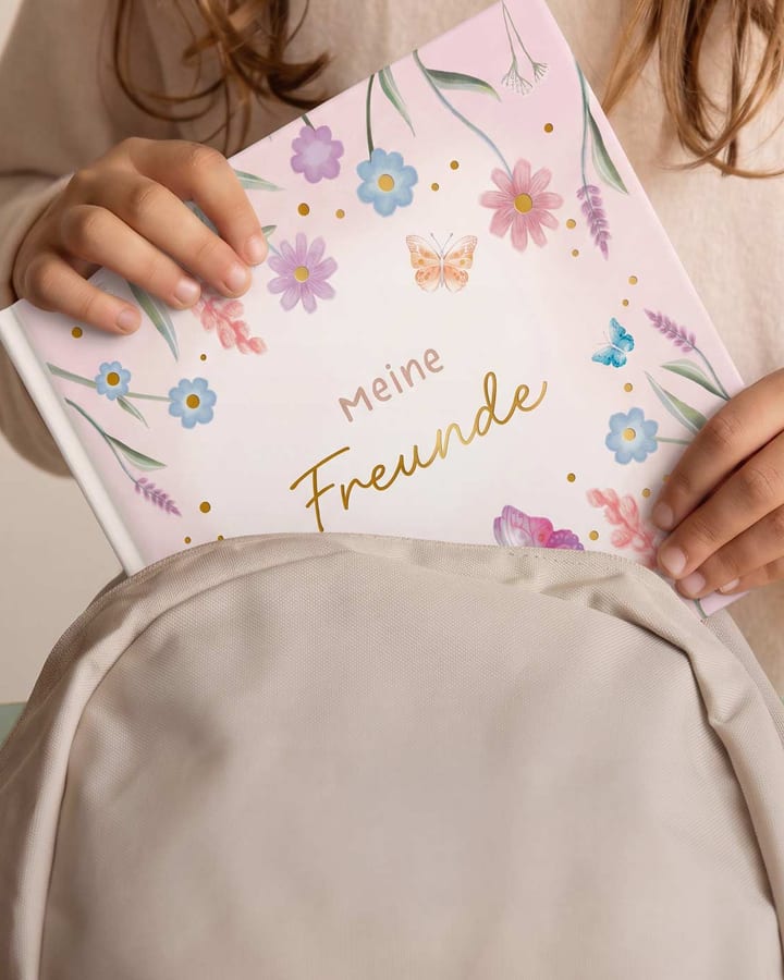 Kinderhände ziehen ein Freundebuch mit Blumenmuster aus einem beigen Rucksack