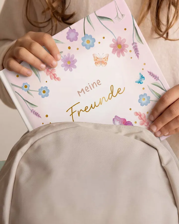 Kinderhände ziehen ein Freundebuch mit Blumenmuster aus einem beigen Rucksack