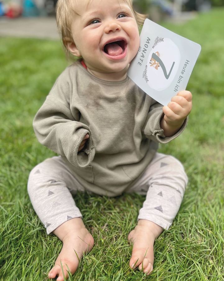 Laughing toddler sitting in the grass holding a Mintkind milestone card