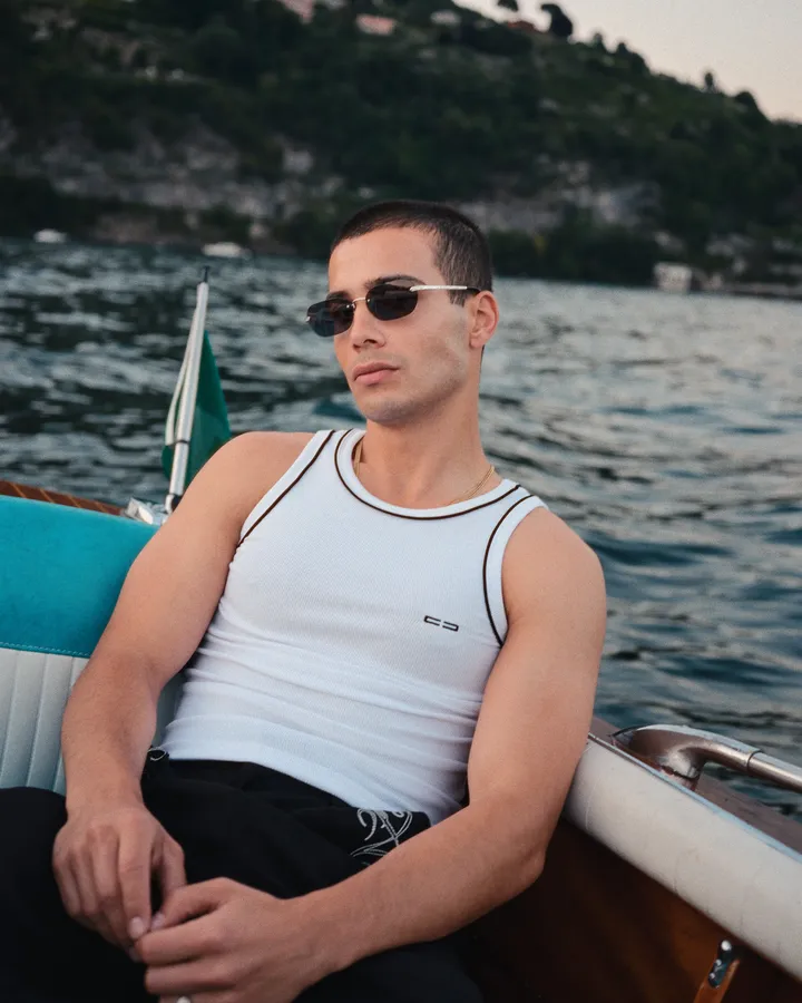 Man in sunglasses and a white tank top sitting in a wooden boat on a lake