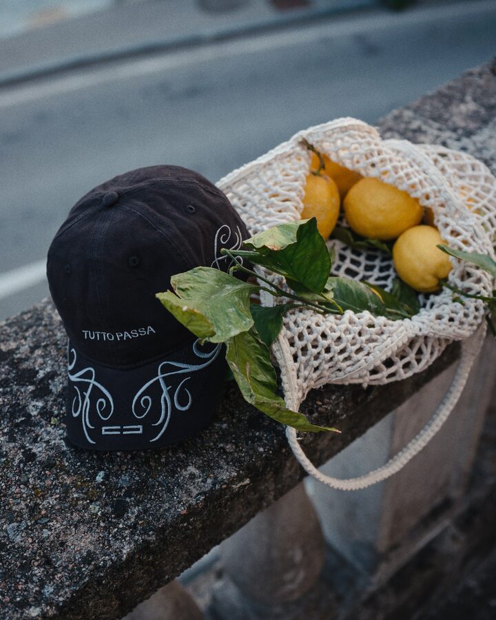 Black embroidered cap next to a net bag of lemons on a stone wall