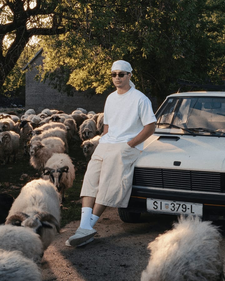 Mann in weissem T-Shirt und heller Shorts lehnt an einem weissen Oldtimer, umgeben von einer Schafherde
