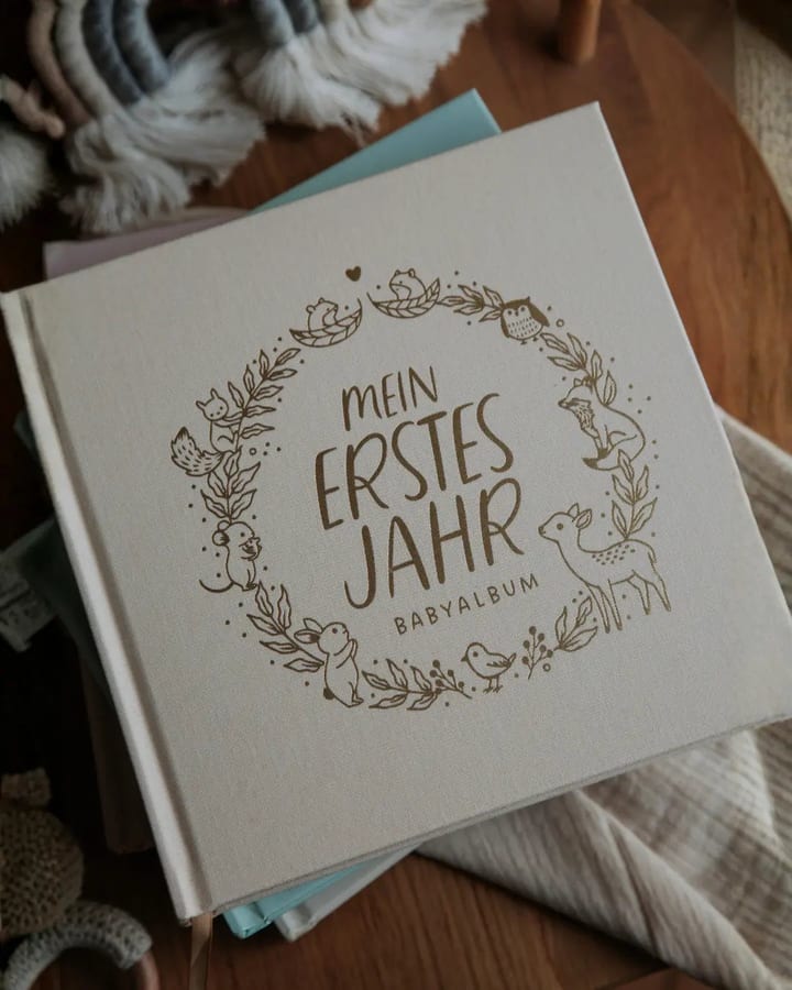 Leinengebundenes Babyalbum mit geprägtem Tierkranz und der Aufschrift Mein erstes Jahr auf einem Holztisch