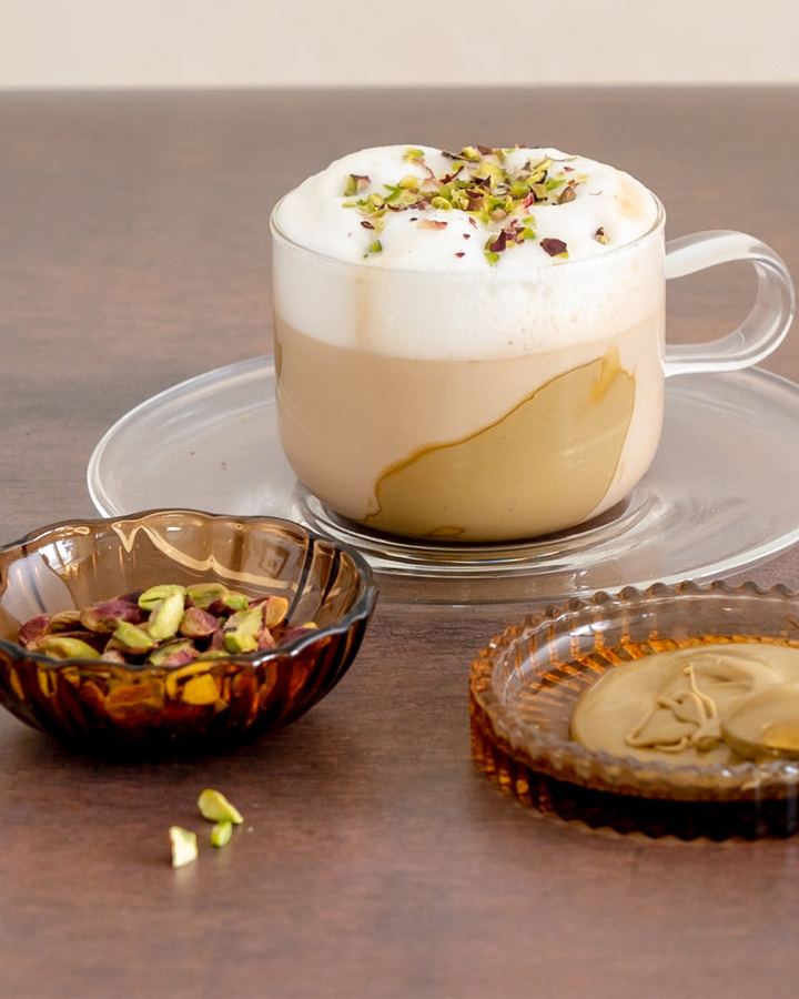 Kaffeespezialität mit Milchschaum und Pistazien im Glas, dazu Schälchen mit Pistaziencreme