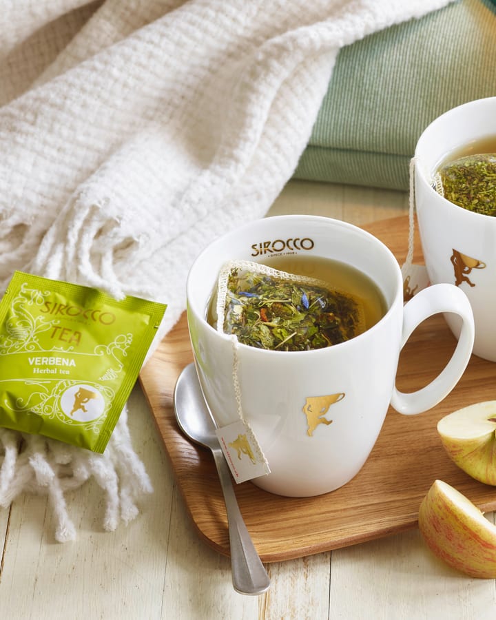 Weisse Sirocco-Tasse mit Kräutertee im Beutel auf Holzbrett, daneben grünes Verbena-Sachet und Apfelschnitze
