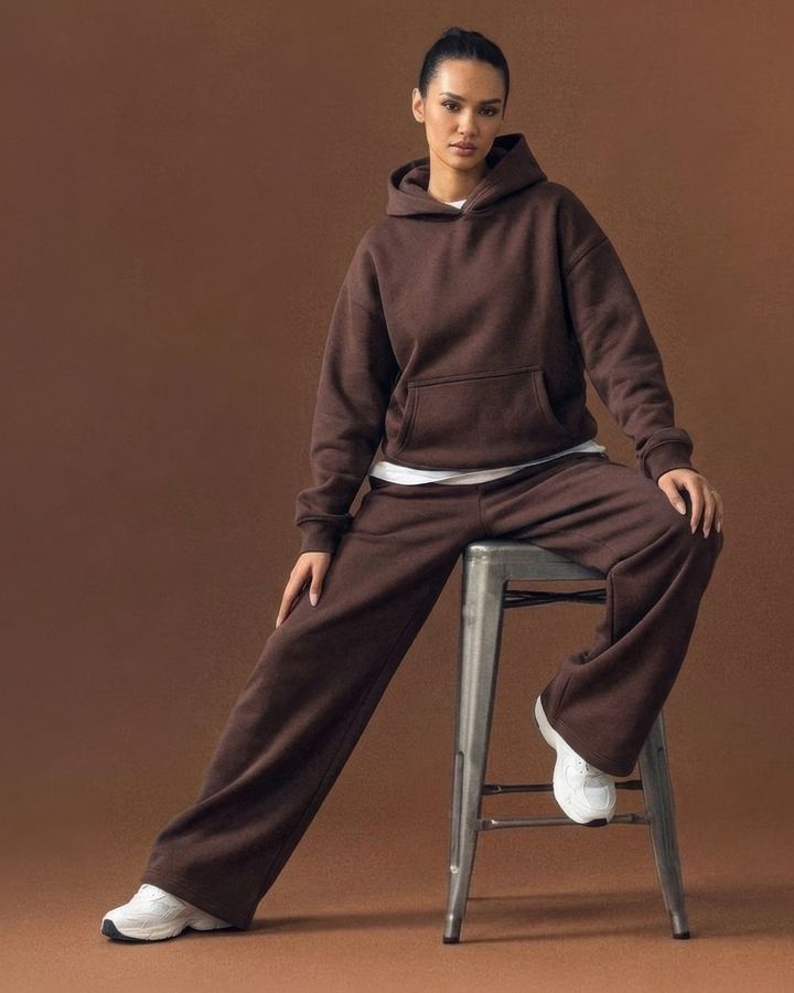 Woman in a brown hoodie and wide sweatpants sitting on a metal stool against a brown backdrop