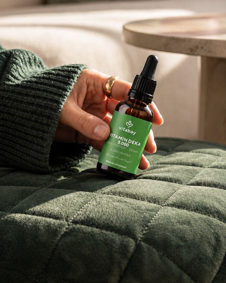 Hand holding a vitamin D dropper bottle above a green quilted throw on a sofa.